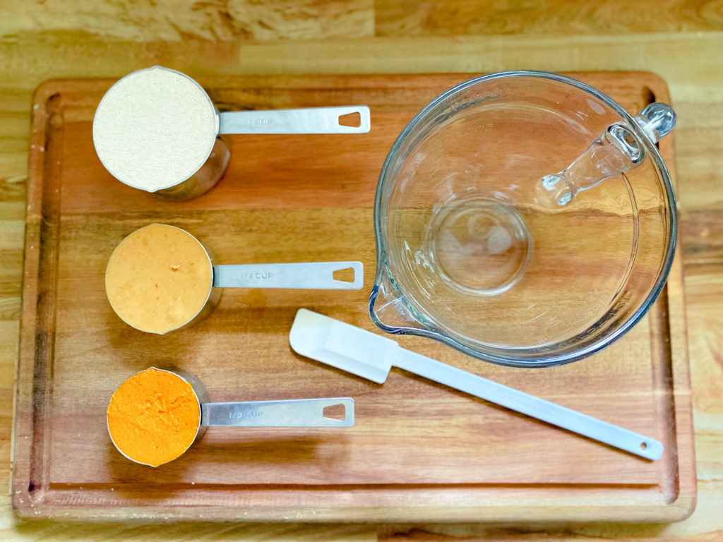 3 ingredients for this dog treat recipe laid out on a wood surface with a mixing bowl and spatula. The flour, peanut butter, and pumpkin puree are divided into separate measuring cups.