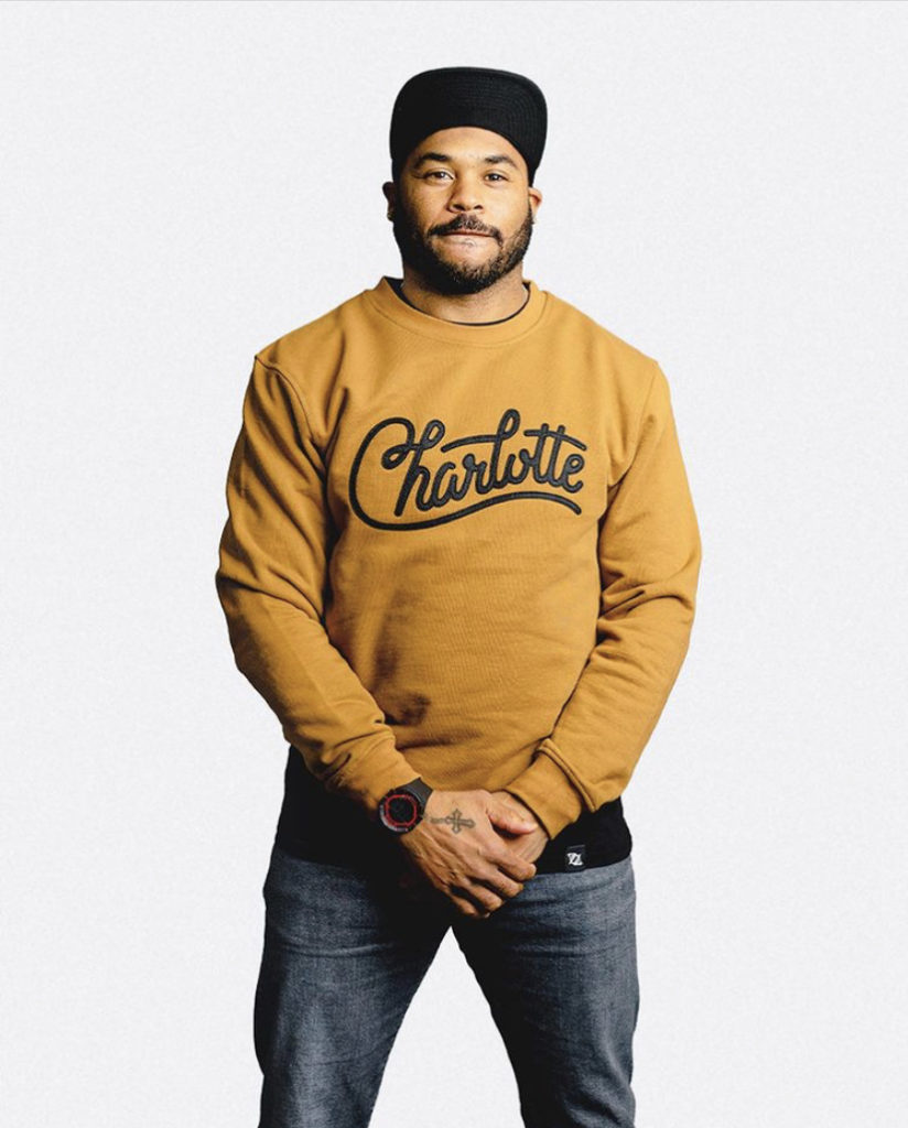 A man wearing a merigold yellow sweatshirt from 704 Shop with the text "Charlotte" on the front in a script lettering