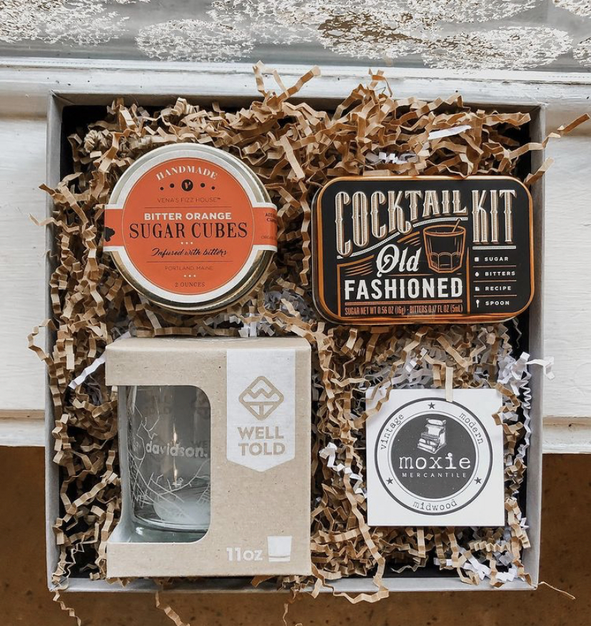 A gift-box featuring sugar cubes, an old-fashioned cocktail kit, and a local whiskey glass set in a box of brown crinkled paper from Moxie Mercantile.