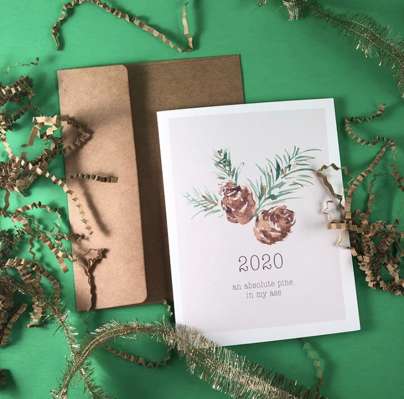 White holiday card with a pinecone on the front and text that says "2020 an absolute pain in my ass" set in front of a brown paper envelope and green background