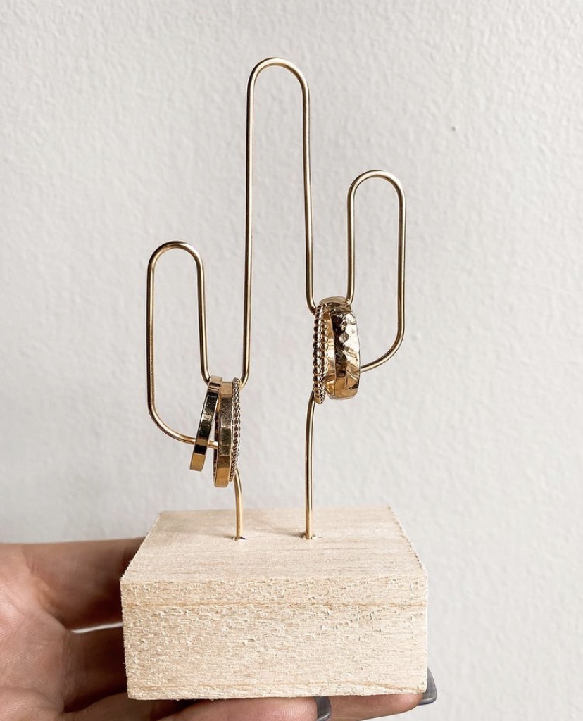 A hand holds a wooden display with a gold wire cactus structure featuring four gold-filled ring designs by Qued Espresso Jewelry