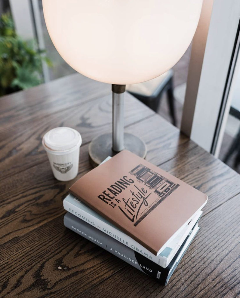 A stack of books, a coffee cup, and a lamp set on a wooden table. The books include a journal, Michelle Obama's "Becoming," and Barak Obama's "A Promised Land"available through Shelves Bookstore