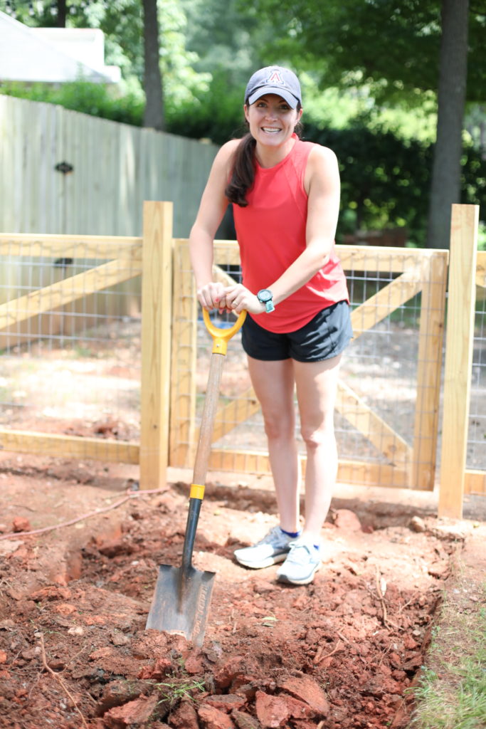 Woman smiling standing on a pile of clay dirt in a red top, black shorts, and navy baseball cap holding a shovel.