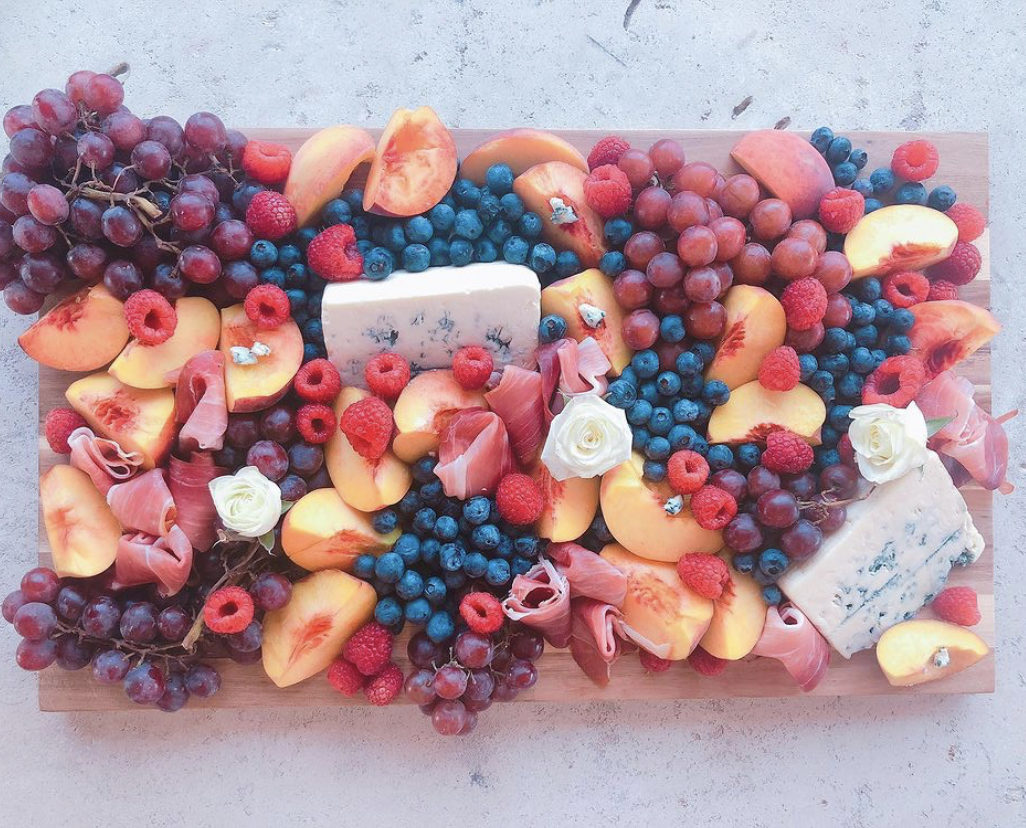 A horizontally-laying meat and cheese board featuring purple grapes, blueberries, blocks of white cheeses, peaches, and raspberries