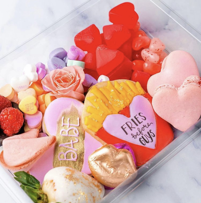 Charcuterie dessert box featuring pink heart-shaped macaroons, a cookie that says "Fries Before Guys" and a pink heart shapped cookie that says "Babe" surround by a pink rose, red heart shaped candies, and conversation heart candies.